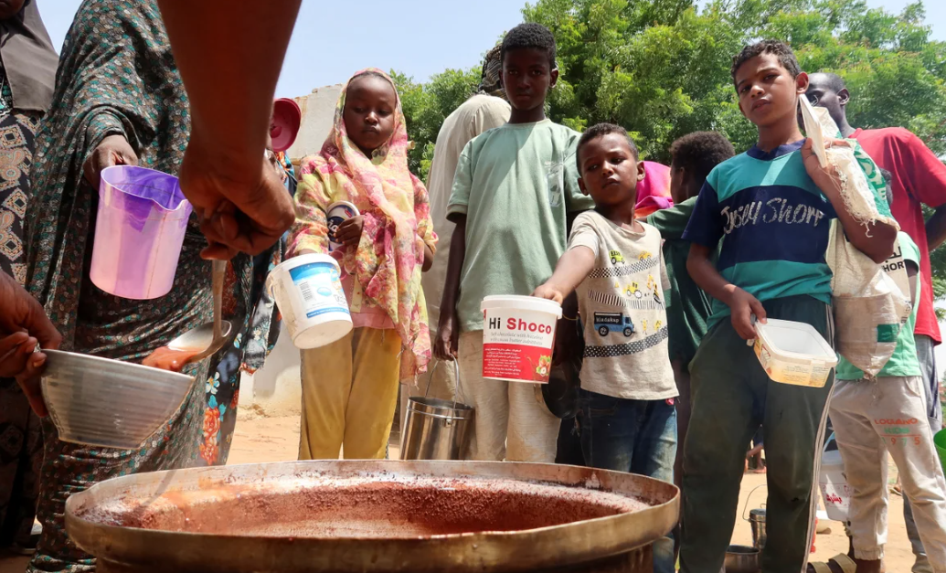 Volunteers in Sudan ladle food from large containers in a community kitchen, providing meals to people affected by war and famine.