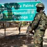 A Mozambican soldier in camouflage carrying a rifle stands beside a road sign near Palma, symbolizing the conflict zone linked to allegations involving TotalEnergies.