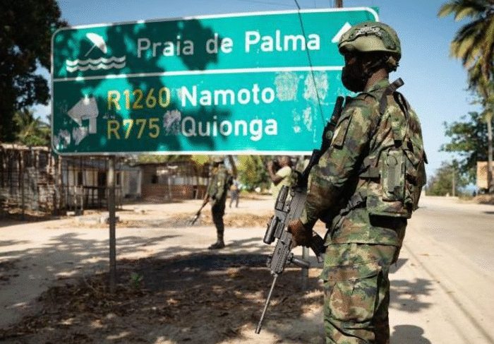 A Mozambican soldier in camouflage carrying a rifle stands beside a road sign near Palma, symbolizing the conflict zone linked to allegations involving TotalEnergies.