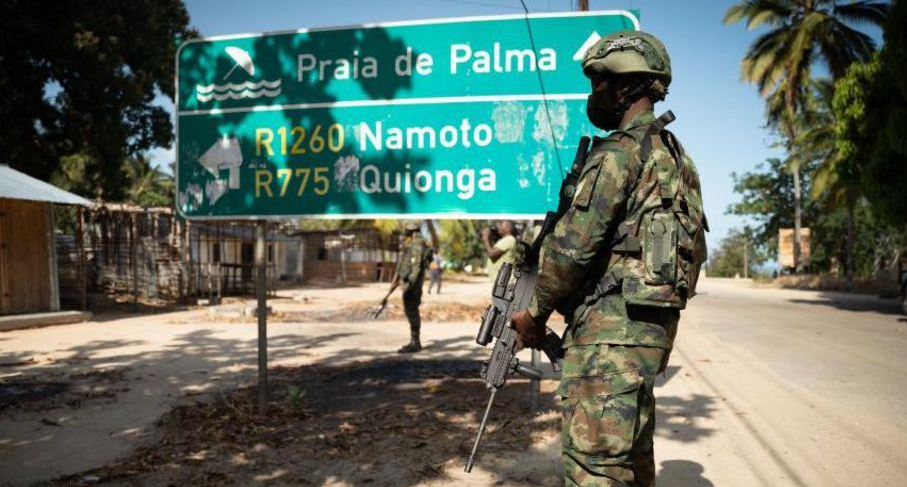 A Mozambican soldier in camouflage carrying a rifle stands beside a road sign near Palma, symbolizing the conflict zone linked to allegations involving TotalEnergies.