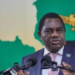 Zambian President Hakainde Hichilema crouching behind a microphone as security personnel move to protect him during a stone-throwing incident at a public rally.