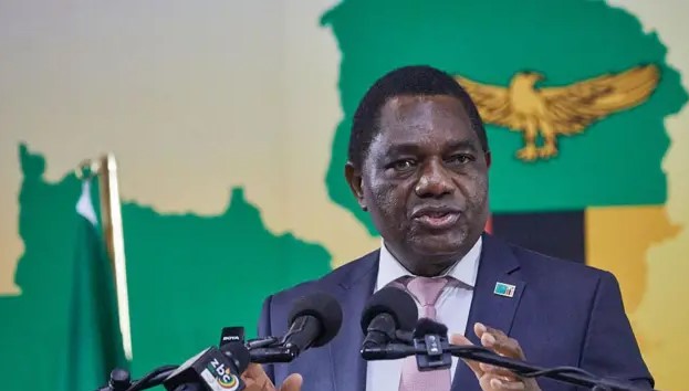 Zambian President Hakainde Hichilema crouching behind a microphone as security personnel move to protect him during a stone-throwing incident at a public rally.