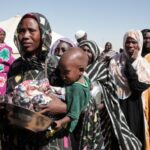 Refugees walking in Sudan with aid tents in the background, illustrating the humanitarian crisis caused by the civil war.